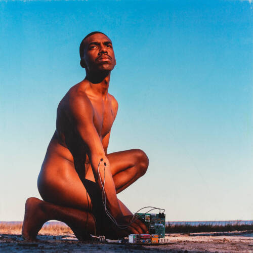 Color photograph of a crouched nude man with wires on his arm and hand.